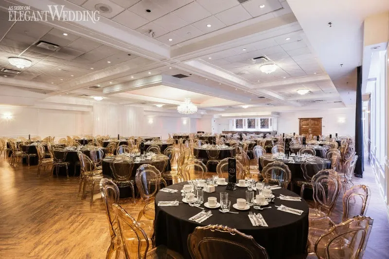 The image showcases a beautifully arranged wedding venue by C’est Classique, categorized under Wedding Venues on Mariage Elegant, Canada. The spacious room features elegantly set round tables with black tablecloths, each adorned with fine china and polished silverware, ready for a wedding reception. The venue is illuminated by opulent chandeliers, which add a touch of sophistication to the ambiance. Transparent chairs provide a modern touch to the classic setting, and the hardwood floors complement the overall elegant atmosphere. The ceiling is decorated with recessed lighting, enhancing the venue’s warm and inviting feel, perfect for a memorable wedding celebration.