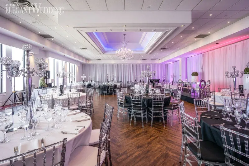 This image showcases a luxurious wedding venue by C’est Classique, categorized under Wedding Venues on Mariage Elegant, Canada. The spacious room features elegantly arranged tables adorned with pristine white tablecloths and neatly folded napkins. Each table is complemented by sophisticated candelabra centerpieces, adding a touch of classic elegance. The ceiling is beautifully adorned with multiple crystal chandeliers, casting a gentle and inviting glow across the room. The walls are draped with soft, white curtains, enhancing the romantic ambiance. Large windows line one side of the venue, allowing natural light to illuminate the space. The setup is ideal for upscale wedding receptions, offering a harmonious blend of elegance and modernity.