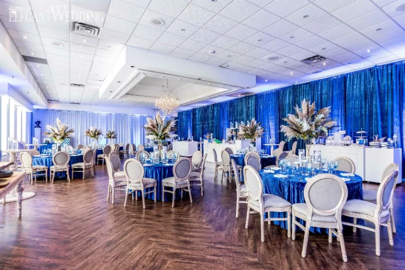 The image showcases a sophisticated wedding venue by C’est Classique, a vendor under the Wedding Venues category on Mariage Elegant, Canada. The setting features elegantly arranged round tables draped in rich blue tablecloths, each adorned with lavish floral centerpieces consisting of white and beige blooms with lush greenery. Chic gray upholstered chairs with curved backs encircle the tables, creating a refined atmosphere. To the side, a grand chandelier hangs from the ceiling, adding a touch of glamour. The room is warmly lit with soft, ambient lighting reflecting off the polished wooden floor, while a backdrop of flowing blue fabric curtains enhances the luxurious ambiance.