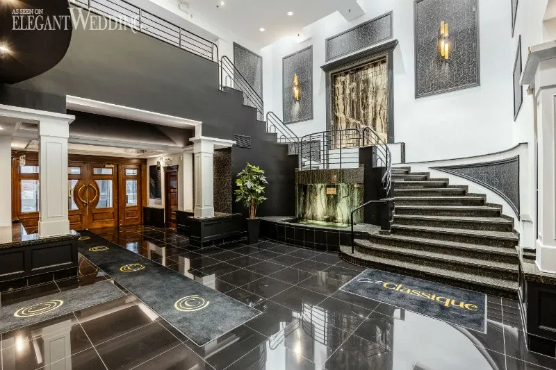 The image showcases the elegant interior of C’est Classique, a premium wedding venue listed on Mariage Elegant in Canada. The venue features a stunning black and white foyer with glossy black tiled flooring and a grand curved staircase, accented with ornate railings. The space is illuminated with modern lighting fixtures that add a sophisticated ambience. A unique waterfall accent is visible near the staircase, enhancing the luxurious atmosphere of this wedding venue. The logo 'C’est Classique' is prominently featured on a rug, reinforcing the venue's branding in a tasteful manner.