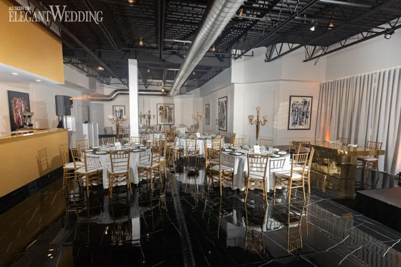 The image showcases the elegant interior of C’est Classique, a prestigious wedding venue under the category of Wedding Venues, featured on Mariage Elegant, Canada. The scene depicts a sophisticated banquet hall with glossy black tiled floors and an industrial-style ceiling. Round tables are neatly arranged and adorned with crisp white tablecloths, complemented by luxurious gold Chiavari chairs. Each table is set with fine dinnerware, and ornate gold candelabras add a touch of opulence. The walls are decorated with modern artwork, enhancing the contemporary and refined atmosphere ideal for hosting memorable wedding receptions.