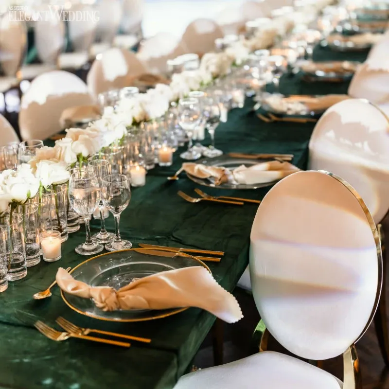 This image showcases an elegant wedding reception setup by vendor C’est Classique, under the wedding venues category. The scene features a long table adorned with a dark green tablecloth, beautifully contrasted by gold-rimmed glassware and neatly folded beige napkins placed on transparent plates. A lush, continuous centerpiece of white roses and greenery runs the length of the table, adding a touch of sophistication. Softly glowing candles are placed intermittently, creating a warm and inviting ambiance. The white chairs, some with gold detailing, perfectly complement the overall luxurious atmosphere, making it an ideal setting for a refined wedding celebration.
