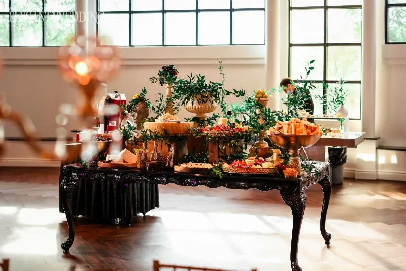 The image showcases an elegantly decorated reception venue offered by C’est Classique, categorized under wedding venues for Mariage Elegant in Canada. A lavishly set buffet table is prominently displayed, featuring an assortment of appetizers and fruit, beautifully arranged in ornate bowls and platters. The table is adorned with lush greenery, adding a touch of natural elegance. Sunlight filters through large windows in the background, illuminating the wooden floors and creating a warm, inviting atmosphere ideal for a wedding celebration.