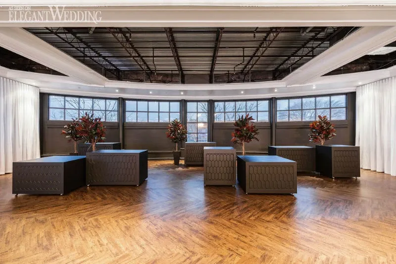 This image showcases the interior of a wedding venue, C’est Classique, categorized under Wedding Venues on Mariage Elegant, Canada. The spacious room features a sleek, modern design with large windows allowing natural light to flood the space. The floor is adorned with elegant herringbone-patterned wood, and dark, textured square tables are strategically placed around the room. Each table is accented with vibrant floral arrangements, adding a touch of color and sophistication to the setting, perfect for wedding celebrations.