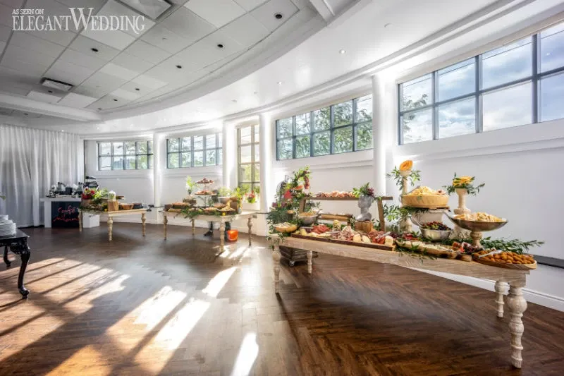 The image showcases a beautifully decorated wedding venue under the vendor C’est Classique, categorized under Wedding Venues for Mariage Elegant in Canada. The room features an elegant, spacious setting with large windows allowing natural light to illuminate the area. Long wooden tables are artfully arranged with a variety of gourmet foods, including fresh fruits, cheeses, and other delicacies, adorned with greenery and floral accents. The hardwood floor and draped white curtains add to the sophisticated and inviting atmosphere, perfect for hosting an elegant wedding reception.