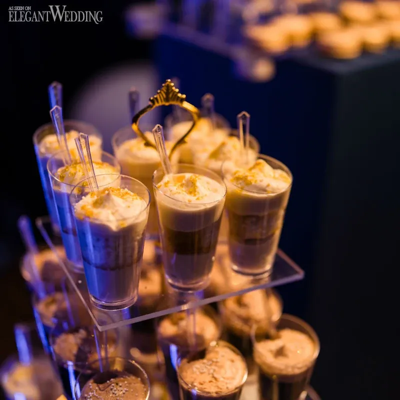 The image showcases a beautifully arranged selection of elegant dessert cups on a tiered glass stand, each topped with a dollop of whipped cream and garnished with sprinkles, under ambient lighting. This setup is part of the exquisite wedding venue decor by C’est Classique, a distinguished vendor in the Wedding Venues category for Mariage Elegant, Canada, highlighting their attention to detail and commitment to creating a luxurious dining experience.