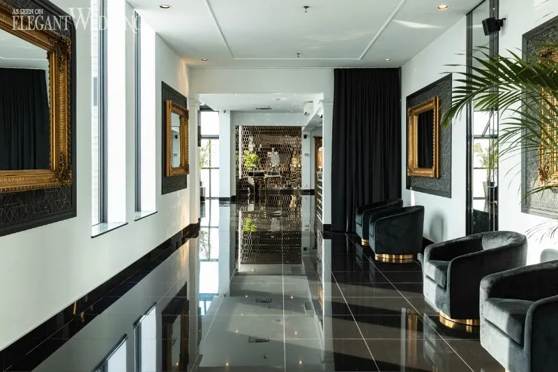 The image showcases a sophisticated and elegant hallway at C’est Classique, a wedding venue listed under the Wedding Venues category on Mariage Elegant, Canada. The space features a glossy black tile floor reflecting the ambient light, with luxurious black velvet chairs neatly aligned along the right wall. Large, ornate gold-framed mirrors adorn the walls, adding a touch of classic elegance. The left side of the hallway is lined with tall windows, allowing natural light to filter through. In the background, there's a glimpse of a stylish bar area, perfect for wedding receptions. The overall ambiance is one of refined luxury, ideal for hosting an upscale wedding event.