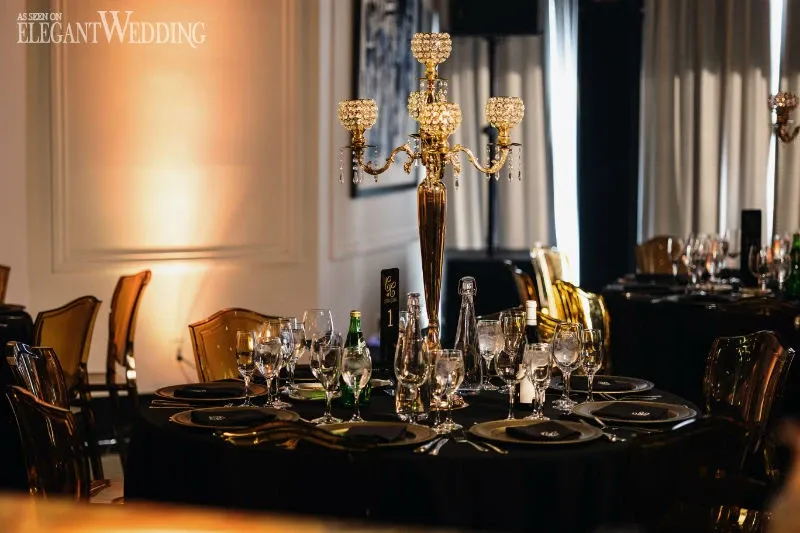 The image showcases a beautifully arranged wedding venue by C’est Classique, categorized under wedding venues. The setting features an elegantly decorated banquet table adorned with a sophisticated gold candelabra as the centerpiece. Each place setting includes polished glassware and neatly arranged black plates, complemented by subtle, warm lighting that creates an intimate and luxurious atmosphere. The background reveals soft drapes and ambient walls, contributing to the elegant and classic theme of the venue. This sophisticated setup highlights C’est Classique's attention to detail, perfect for those seeking a refined wedding experience in Canada.