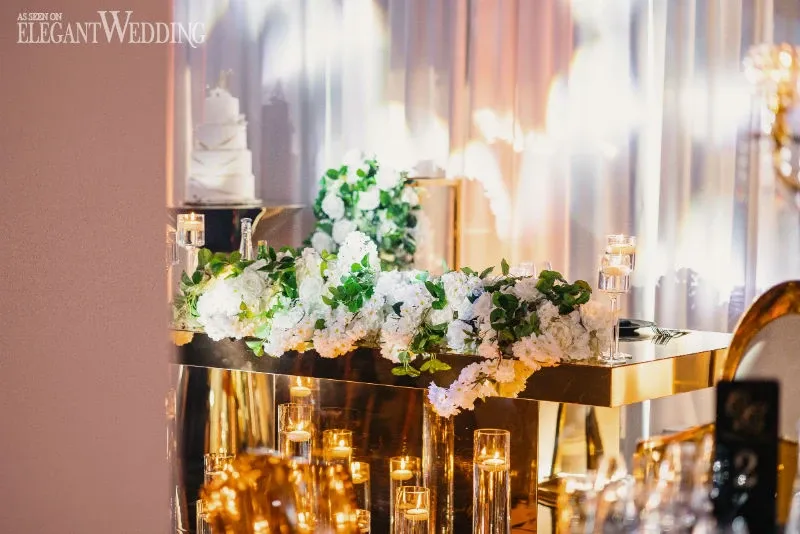 The image showcases a beautifully decorated wedding venue by C’est Classique, categorized under Wedding Venues on Mariage Elegant, Canada. The elegant setup features a luxurious metallic table adorned with lush white and green floral arrangements. A tiered wedding cake, subtly positioned in the background, adds to the celebratory ambiance. The table is surrounded by soft candlelight, casting a warm and inviting glow throughout the space.