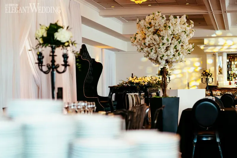 The image showcases a beautifully decorated wedding venue by C’est Classique, falling under the wedding venues category. The setting features elegant black chairs and a grand black armchair, contributing to a sophisticated atmosphere. A stunning floral arrangement of white blossoms sits prominently, complementing the elegant and romantic decor. The room is warmly lit, with soft lighting highlighting a lavish display of white and yellow flowers on a long table. The refined details and luxurious ambiance make it an ideal choice for weddings in Canada.