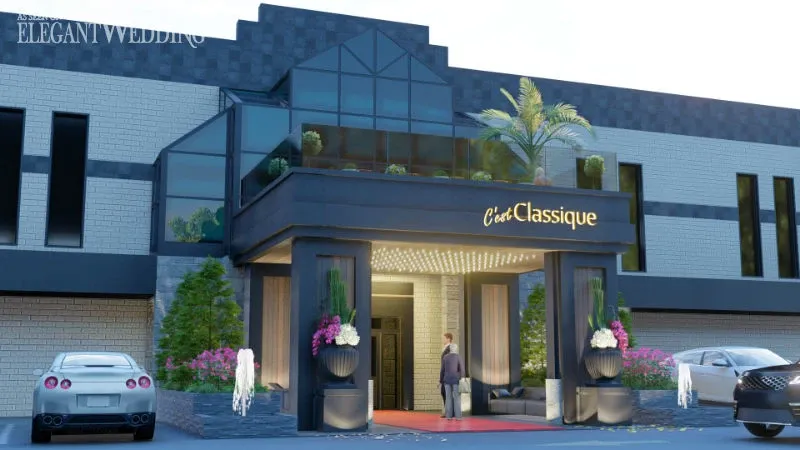 The image showcases C’est Classique, a premier wedding venue featured on Mariage Elegant, Canada. The venue's entrance is elegantly designed with a modern facade, adorned with large glass panels and palm trees, creating a welcoming and sophisticated atmosphere. A person stands by the entrance on a vibrant red carpet, which leads into the building, emphasizing the venue’s luxurious appeal. Flanking the entrance are lush green plants in stylish planters, adding a touch of natural beauty to the setting. A sleek sports car is parked nearby, enhancing the sense of opulence associated with this wedding venue.