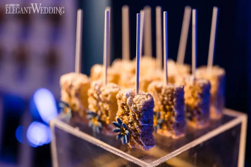 The image features an elegant display of gourmet treats on sticks, resembling rice crispy pops, arranged neatly on a clear acrylic stand. Each treat is adorned with a small, decorative flower, adding a touch of sophistication suitable for upscale celebrations. The ambient lighting casts a warm glow, enhancing the luxurious atmosphere typical of C’est Classique, a featured wedding venue vendor in the wedding planning category on Mariage Elegant, Canada.