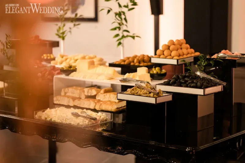 The image showcases an elegant buffet setup by C’est Classique, a renowned wedding venue in Canada, under the category of Wedding Venues. The spread features a stylish arrangement of gourmet cheeses, assorted breads, and olives, beautifully presented on sleek black stands. The sophisticated display is complemented by leafy green accents, providing a touch of natural elegance. With soft lighting enhancing the warm, inviting atmosphere, this image embodies the refined and luxurious offerings available for wedding celebrations at C’est Classique.