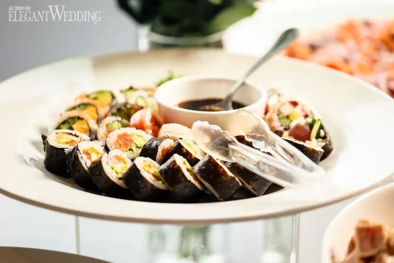 This image showcases a beautifully arranged platter of assorted sushi rolls, featuring a variety of fillings such as fresh vegetables and fish, elegantly displayed at a wedding venue by C’est Classique. The sushi is artfully presented in a circular formation on a white platter, accompanied by a small bowl of soy sauce and clear serving tongs. The luxurious presentation reflects the sophistication of the wedding venue category offered by C’est Classique, highlighting their attention to detail and culinary excellence for elegant weddings.