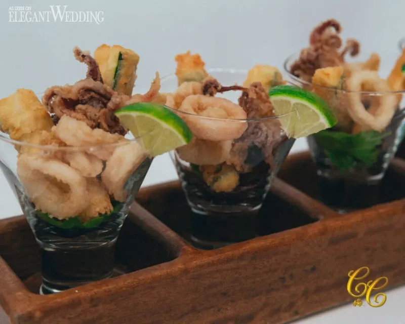 The image showcases a beautifully presented appetizer display from C’est Classique, part of the wedding venues category on Mariage Elegant, Canada. Three elegant glass cups are filled with crispy calamari garnished with slices of lime. Each cup sits in a wooden tray, making it a stylish choice for a sophisticated wedding reception. The presentation emphasizes quality and attention to detail, suitable for a high-end wedding venue.