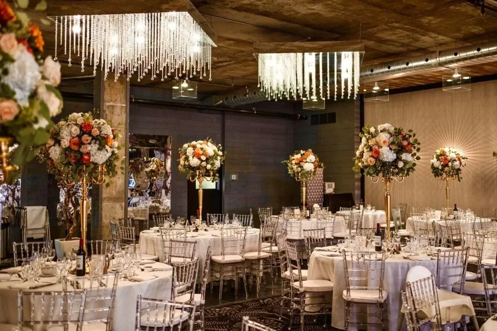 The image showcases a beautifully decorated wedding venue by The PMG Group, categorized under wedding venues, featured on Mariage Elegant in Canada. The setting includes elegantly arranged round tables draped in white linens, each adorned with tall floral centerpieces consisting of vibrant roses and assorted flowers. Transparent chiavari chairs surround the tables, exuding a modern yet classic elegance. Overhead, luxurious chandeliers with cascading crystals add a touch of glamour and sophistication to the venue. The space is warmly lit, highlighting the rustic charm of the exposed brick and wooden elements, creating an inviting and romantic atmosphere for a wedding celebration.