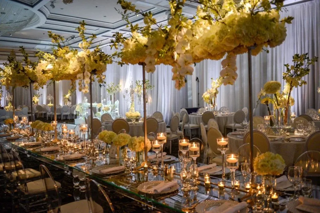 The image showcases an elegant wedding venue managed by The PMG Group, under the category of Wedding Venues, for Mariage Elegant in Canada. The venue features a beautifully decorated banquet hall with lavishly set tables adorned with crystal glassware, fine china, and an array of lit candles creating a warm and inviting atmosphere. Lush floral arrangements, predominantly in shades of yellow and white, elegantly hang overhead, adding a touch of sophistication. The backdrop includes tall, sheer drapery, enhancing the room's grandeur, while round tables and plush seating accommodate guests, promising a memorable wedding celebration.