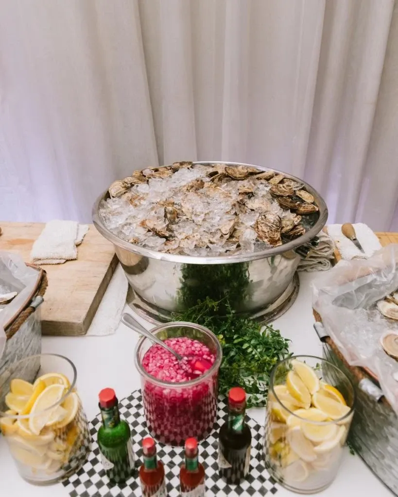 The image features an elegant wedding setup by The PMG Group, categorized under Wedding Venues for Mariage Elegant in Canada. A polished metal bowl is centrally placed, filled with ice and topped with opened oysters, indicating a luxurious seafood offering. Surrounding the bowl are containers with lemon wedges, bottles of sauce, and a jar containing chopped onions, adding color and variety to the display. The backdrop consists of a white curtain, suggesting a sophisticated and serene wedding environment. This scene is indicative of a high-quality shellfish station, often found at upscale wedding receptions.