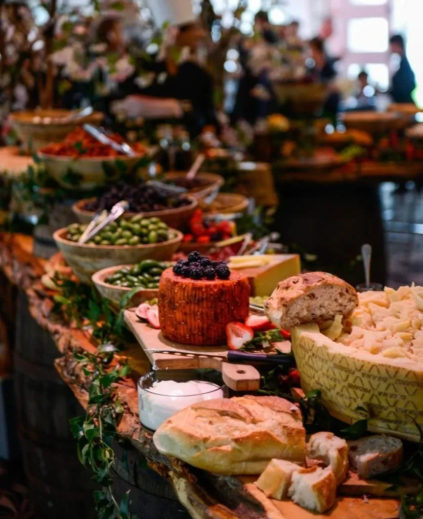 The image shows an elegantly arranged wedding buffet setup at The PMG Group, a wedding venue in the Mariage Elegant directory, Canada. A rustic wooden table is adorned with an assortment of artisanal cheeses, fresh fruits, and loaves of crusty bread. A focal point of the display is a round cheese wheel decorated with a cluster of blueberries and surrounded by figs and leaves for a natural touch. The background features lush greenery and softly blurred silhouettes of guests, creating an inviting and sophisticated atmosphere perfect for a wedding celebration.