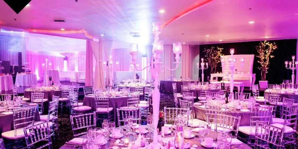 The image showcases a beautifully arranged wedding venue by The PMG Group in the Wedding Venues category, featuring an elegantly decorated banquet hall. The hall is adorned with round tables covered in white tablecloths and surrounded by transparent Chiavari chairs. Each table is meticulously set with glassware, cutlery, and floral centerpieces. The venue is illuminated with soft, ambient lighting in hues of pink and purple, creating a romantic atmosphere. Candelabras and decorative drapery add to the sophisticated decor, highlighting the attention to detail ideal for a wedding celebration.