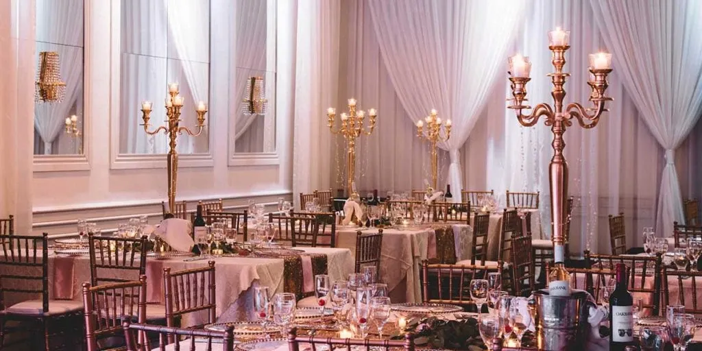 The image showcases a beautifully decorated wedding venue by The PMG Group, categorized under Wedding Venues. The elegant space features round tables adorned with white tablecloths and fine glassware, surrounded by gold Chiavari chairs. Tall, ornate golden candelabras with lit candles are prominently placed on each table, enhancing the romantic atmosphere. The backdrop consists of soft, white drapery and large mirrors, completing the sophisticated and luxurious setting suitable for an upscale wedding event.