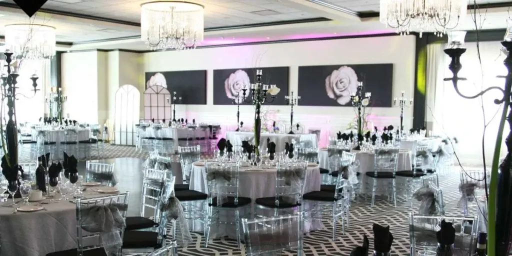 The image displays a beautifully arranged wedding venue by The PMG Group, categorized under wedding venues. The setting features elegantly decorated tables with white tablecloths and black napkins, surrounded by transparent chairs. Luxurious chandeliers hang from the ceiling, casting a soft glow across the room. The walls are adorned with large, artistic rose paintings, adding a romantic touch. The sophisticated decor and ambient lighting make it an ideal setting for an upscale wedding reception, perfectly capturing the essence of a stylish and memorable celebration in Canada.