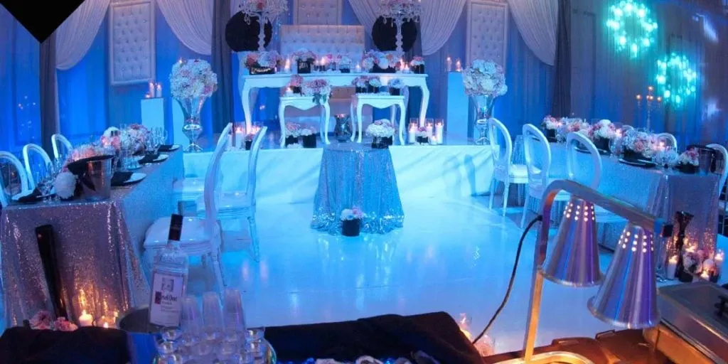 The image showcases an elegant wedding venue by The PMG Group in the Wedding Venues category. The setting features a beautifully decorated reception area with shimmering blue and white drapery adorning the walls and ceiling. The centerpiece of the venue is a sophisticated table arrangement, illuminated by soft, ambient lighting, and surrounded by transparent chairs. Tables are adorned with sparkling sequin tablecloths and tasteful floral arrangements in pastel hues, adding a touch of romance. The overall atmosphere is one of opulence and elegance, perfect for a memorable wedding celebration.