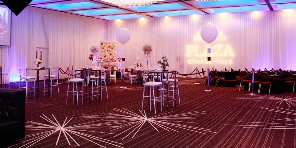 The image showcases an elegantly decorated wedding venue by The PMG Group, categorized under Wedding Venues on Mariage Elegant, Canada. The spacious room features high ceilings with modern, geometric lighting fixtures casting a warm glow. The area is adorned with tall cocktail tables surrounded by transparent chairs, each table topped with floral centerpieces. In the background, a decorative display with soft pink and white flowers adds a touch of romance. The ambiance is enhanced by soft purple lighting along the walls, creating an inviting atmosphere perfect for a wedding reception.