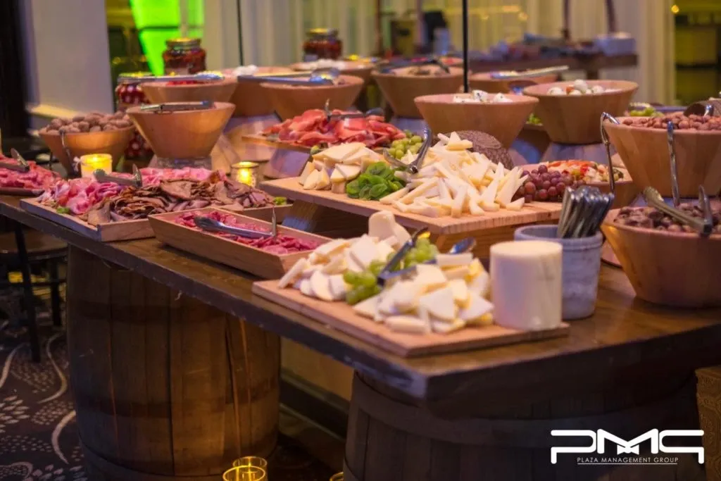 The image features a beautifully arranged charcuterie spread at a wedding venue provided by The PMG Group, a vendor in the wedding venues category. Several wooden boards display a variety of meats, cheeses, fresh fruits, and vegetables, artfully presented on a rustic wooden table supported by barrels, adding a charming touch to the event atmosphere. Decorative candles glow softly around the setup, creating a warm and inviting ambiance suitable for a wedding celebration. The background captures a glimpse of elegant dining settings, reinforcing the sophistication of the venue.
