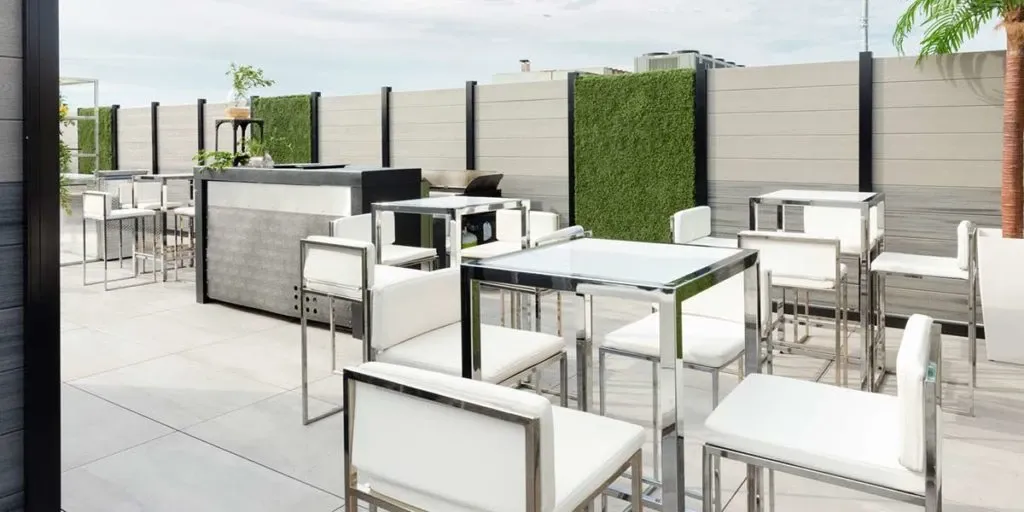 The image showcases a stylish outdoor setup offered by The PMG Group, categorized under Wedding Venues for Mariage Elegant in Canada. The scene features modern, sleek furniture with several high tables and square chairs, all with metallic frames and white cushions, creating a sophisticated ambiance. The venue's perimeter is enclosed by contemporary fencing, adorned with lush green vertical garden panels, adding a touch of nature. A well-equipped bar is visible, hinting at a space tailored for elegant wedding receptions and gatherings.