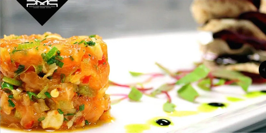 The image features a beautifully presented dish of vibrant orange tartare garnished with finely chopped herbs, showcasing the culinary elegance of The PMG Group, a vendor in the Wedding Venues category. This artistic plating includes delicate drizzles of sauce on a pristine white plate, with a blurred background highlighting other components of the appetizer. The sophisticated presentation reflects the upscale dining experience that The PMG Group offers for wedding events, set against the branding visible in the corner, reinforcing the vendor's identity.