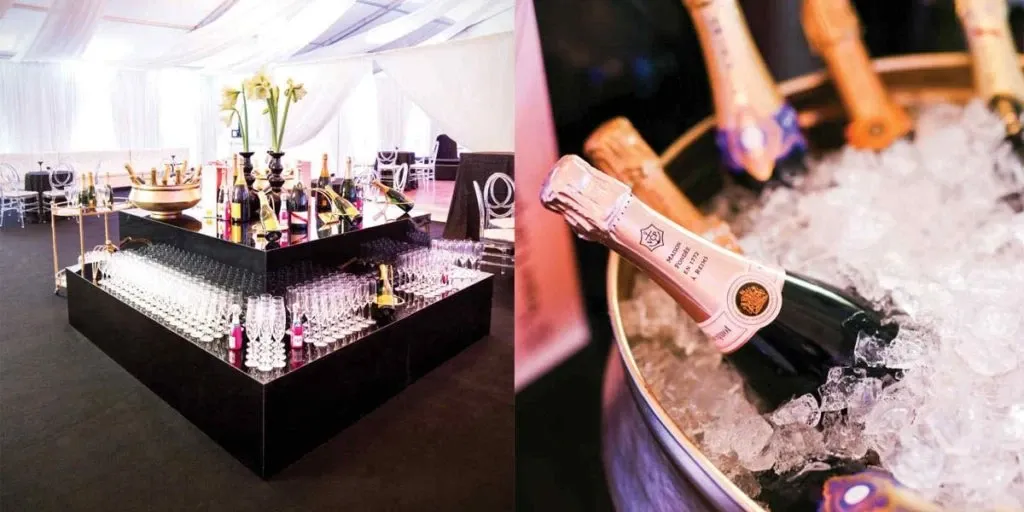 The image showcases an elegant wedding reception venue by The PMG Group, categorized under Wedding Venues. On the left, a stylish bar setup features a sleek, black square counter adorned with numerous champagne bottles and rows of neatly arranged glasses, ready for toasting. White drapes hang from the ceiling, adding a touch of sophistication to the venue. On the right, a close-up image reveals a bucket filled with ice, housing champagne bottles, indicating the venue's readiness to cater to a classy wedding celebration. This setup highlights The PMG Group's commitment to providing a refined and luxurious atmosphere for wedding events.
