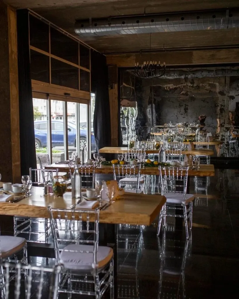 The image showcases an elegant wedding venue by The PMG Group, categorized under Wedding Venues, featured on Mariage Elegant, Canada. The setting includes a series of long wooden tables adorned with crisp white table settings, silver cutlery, and clear glasses, creating a refined atmosphere. Transparent chairs add a modern touch, while the softly lit room features large windows that bring in natural light, enhancing the chic décor. Reflections from the polished floor and the industrial-style ceiling, with exposed ducts, contribute to the venue's sophisticated and contemporary design, ideal for a memorable wedding celebration.