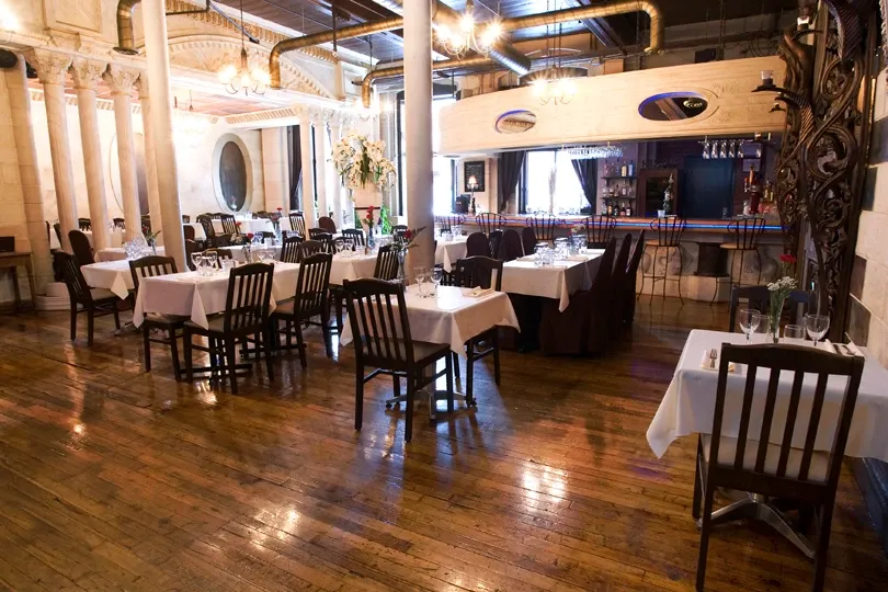 The image showcases the interior of L'Ambroisie et L'Espace Canal, a wedding venue listed under Mariage Elegant in Canada. The venue features elegantly set tables draped in white linens, arranged across a polished hardwood floor. The atmosphere is warm and inviting, with soft lighting accented by ornate ceiling details and columns. A well-stocked bar is visible in the background, adding to the venue's sophisticated charm. This setting is ideal for hosting memorable wedding receptions in a stylish and intimate environment.