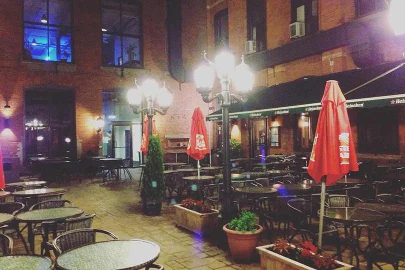 This image showcases the charming courtyard of L'Ambroisie et L'Espace Canal, a premier wedding venue in Canada. The setting features a cobblestone patio adorned with round tables and chairs, perfect for outdoor receptions or gatherings. Brightly lit vintage-style street lamps add a romantic ambiance, while red umbrellas provide a touch of color and shade for daytime events. The backdrop of rustic brick walls enhances the venue's elegant yet inviting atmosphere, ideal for weddings and celebrations.
