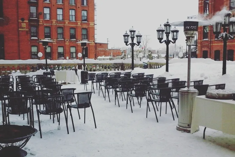 The image shows an outdoor wedding venue at L'Ambroisie et L'Espace Canal, part of the wedding venues category on Mariage Elegant, Canada. The scene is set on a snowy day, with rows of black metal chairs arranged for a wedding ceremony. Several vintage-style lampposts line the area, adding a touch of elegance. Tents cover tables to the side, suggesting a setup for dining or reception. The red brick building in the background, with its classic architectural features, provides a charming and historic backdrop for the venue.