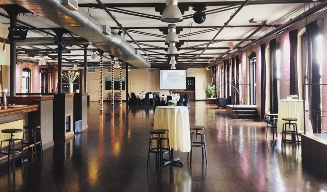The image shows an elegant wedding venue by L'Ambroisie et L'Espace Canal, categorized under Wedding Venues. The spacious interior features a polished wooden floor and exposed industrial-style ceiling beams with suspended lighting. Tall windows line the side walls, allowing natural light to flood the space. The room is set up with high tables draped in white tablecloths, perfect for wedding receptions or cocktail hours. A projector screen is visible at the end of the room, suggesting facilities for presentations or slideshows. The venue exudes a warm ambiance ideal for weddings or special events.