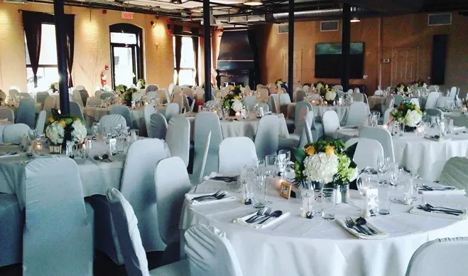 The image showcases a beautifully arranged wedding venue at L'Ambroisie et L'Espace Canal, highlighting its elegant setting perfect for weddings. The room is filled with round tables draped in white linens, each adorned with tasteful floral centerpieces featuring white and green blooms. Chairs are covered with white fabric, creating a cohesive and sophisticated ambiance. Soft lighting complements the exposed brick walls and industrial ceiling, adding to the charm of this wedding venue. The arrangement suggests an intimate and stylish atmosphere for wedding celebrations.
