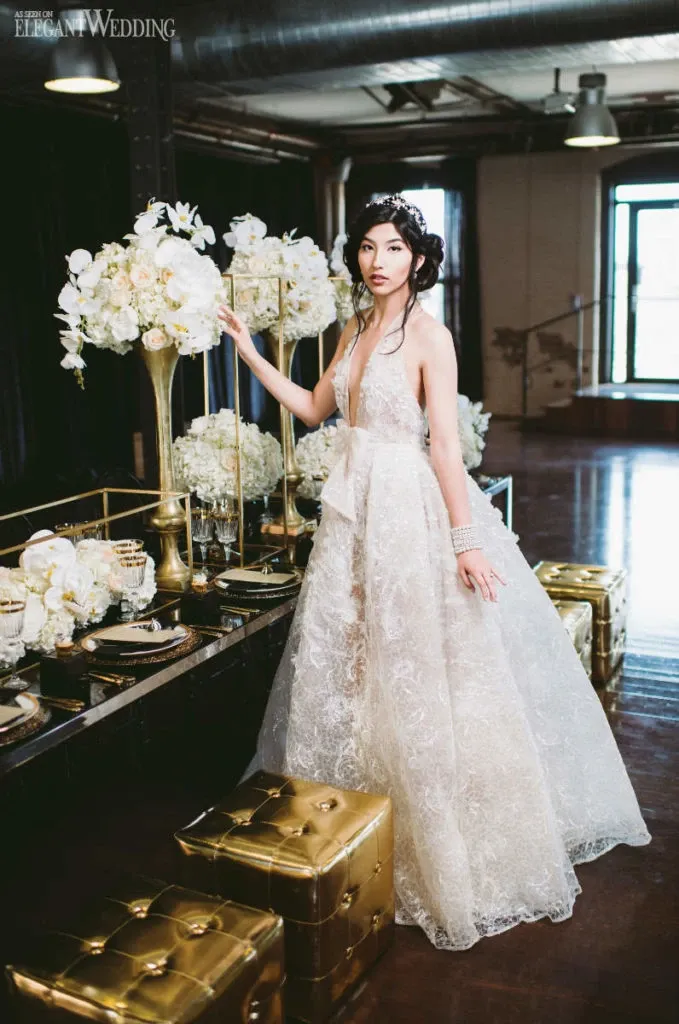 The image showcases a bride standing elegantly beside a lavishly decorated wedding reception table at L'Ambroisie et L'Espace Canal, a wedding venue in Canada. The table is adorned with luxurious gold accents, tall floral arrangements of white flowers, and an elegant centerpiece. The bride is wearing a stunning lace wedding gown with intricate details, and her hair is styled in an elegant updo, embodying a classic yet modern bridal look. The venue's spacious interior with exposed beams and large windows adds a touch of industrial elegance to the setting. This scene perfectly captures the essence of a sophisticated and elegant wedding celebration.