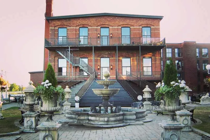 The image showcases L'Ambroisie et L'Espace Canal, a premier wedding venue in Canada, under the category of Wedding Venues. The scene features a charming brick building with large windows, complemented by a grand outdoor staircase leading to the entrance. In the foreground, there is an elegant stone fountain surrounded by decorative planters filled with lush greenery and white flowers. This picturesque setting is ideal for weddings, offering both historical charm and a romantic ambiance for special celebrations.