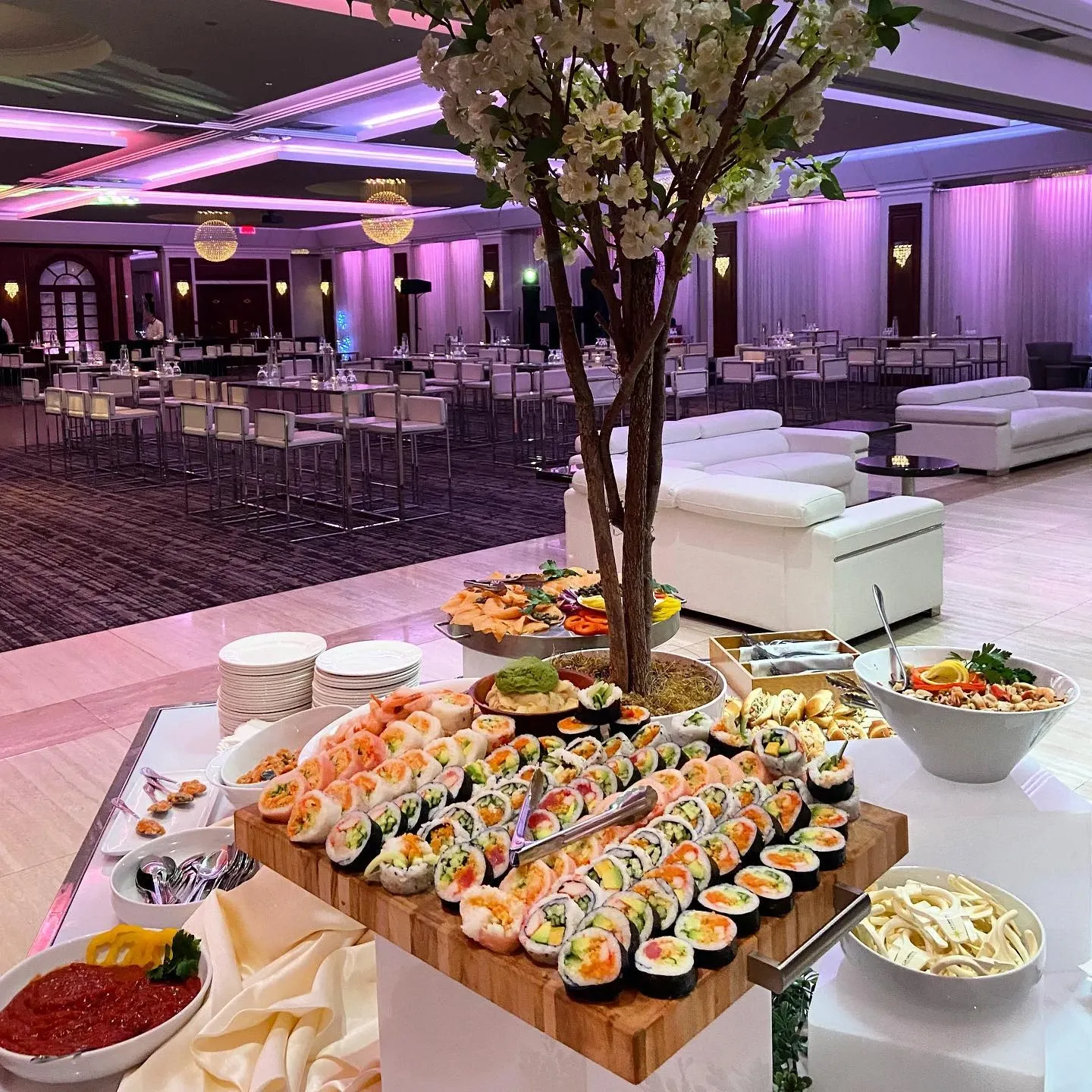 The image shows an elegant wedding venue, Le Crystal, in Canada, showcasing its catering services in a beautifully set event space. A luxurious arrangement of sushi and other gourmet dishes is displayed prominently on a table in the foreground, surrounded by plates and decorative elements. In the background, the venue features a spacious room with white draped walls, modern chandeliers, and ambient purple lighting, creating an inviting atmosphere. Comfortable white lounge seating and tall cocktail tables are arranged throughout the space, perfect for guests to mingle during a wedding reception. This image highlights Le Crystal’s sophisticated wedding venue offerings ideal for elegant celebrations.