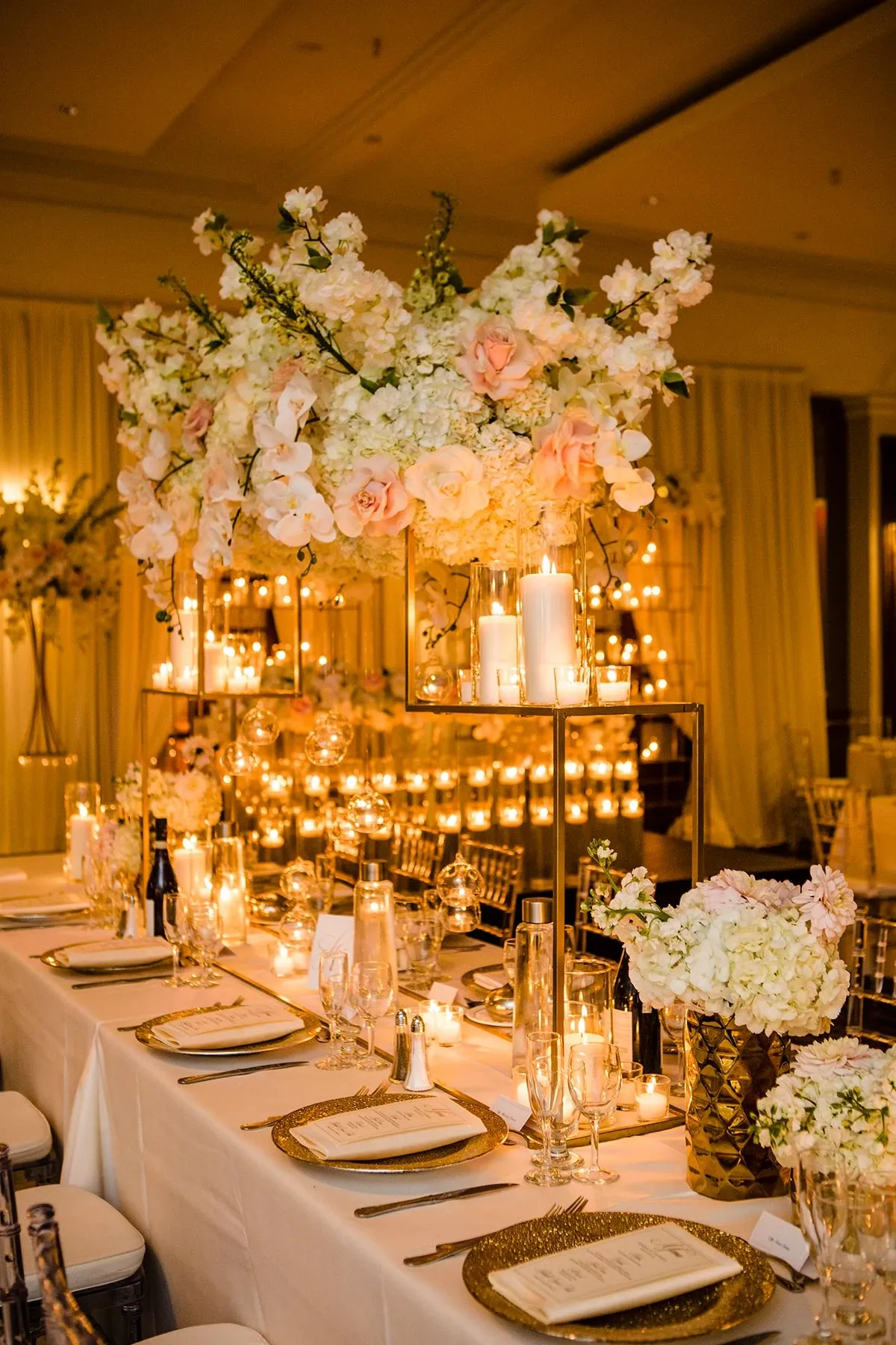 The image showcases an elegant wedding reception setup at Le Crystal, a premier wedding venue in Canada. The table is adorned with luxurious floral arrangements featuring white and blush pink flowers elegantly set in tall, gold stands. The soft, ambient glow from numerous candles enhances the romantic atmosphere. The table is neatly arranged with sparkling glassware, gold-rimmed plates, and exquisite, personalized menus, embodying the sophisticated and opulent style expected from a top-tier wedding venue like Le Crystal.