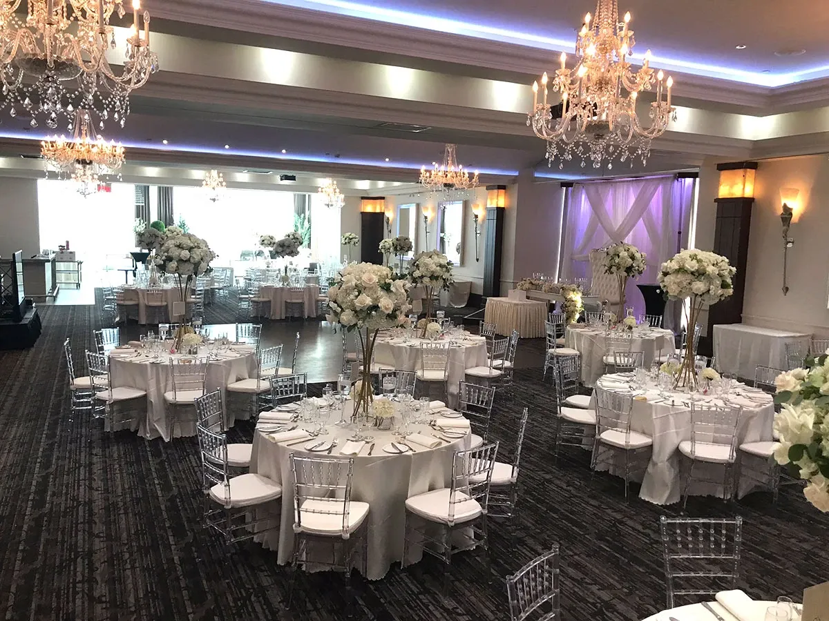 This image showcases the interior of Le Crystal, a top choice for wedding venues listed on Mariage Elegant, Canada. The venue features an elegantly decorated banquet hall with large, sparkling chandeliers illuminating the space. Round tables are meticulously arranged, each adorned with white tablecloths and surrounded by clear, modern chairs. The tables are beautifully set with fine china, glassware, and tall floral centerpieces featuring white and cream flowers. In the background, a softly lit area appears ideal for a wedding ceremony or reception. The ambiance is luxurious and sophisticated, perfect for an unforgettable wedding celebration.