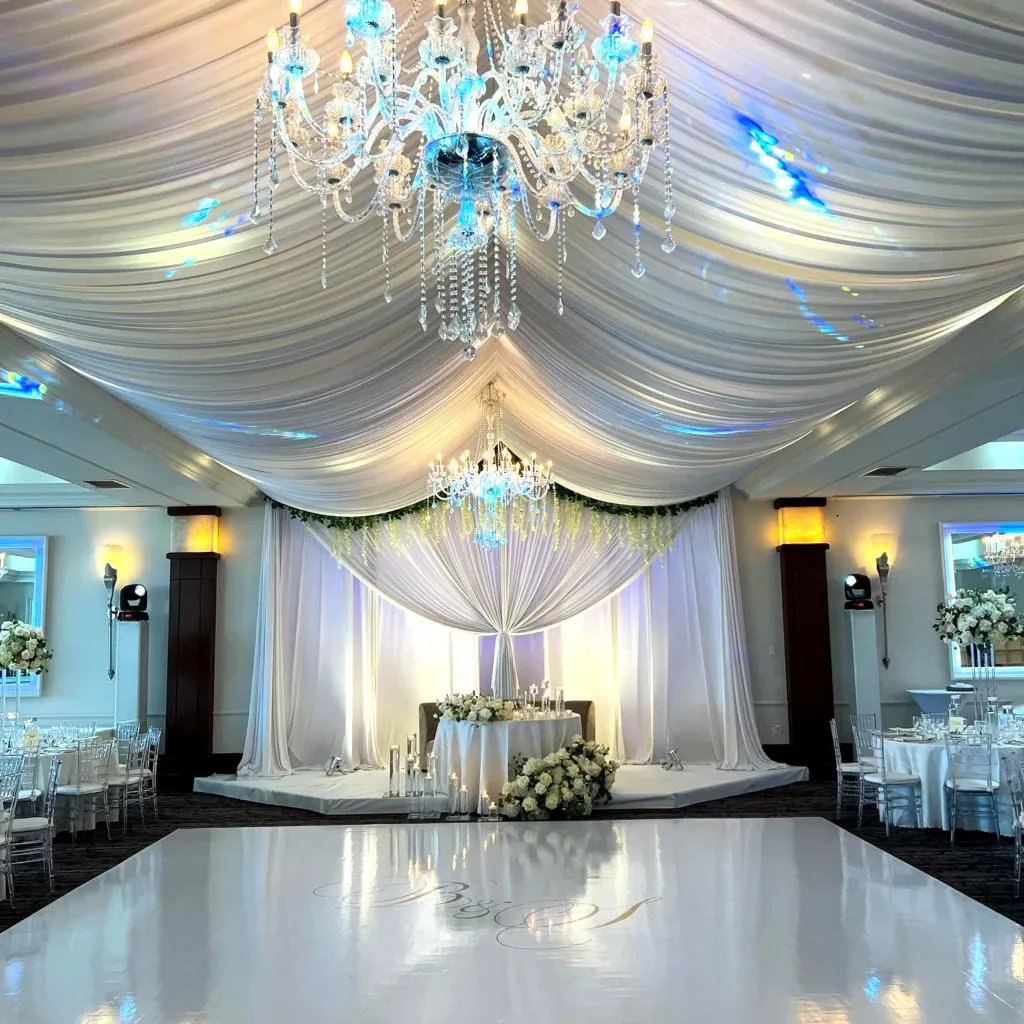 This image showcases Le Crystal, a premier wedding venue in the wedding planning and vendor listing site, Mariage Elegant, Canada. The venue features an elegantly draped ceiling with luxurious white fabric and two stunning crystal chandeliers that cast a soft, ambient glow. The centerpiece of the room is a beautifully decorated sweetheart table adorned with floral arrangements, set against a backdrop of sheer white curtains accented with greenery. The polished white dance floor in the foreground is impeccably maintained, perfect for a memorable first dance. The setting is complete with elegantly arranged tables and chairs, each adorned with sophisticated floral centerpieces, creating a luxurious and romantic atmosphere ideal for a wedding celebration.