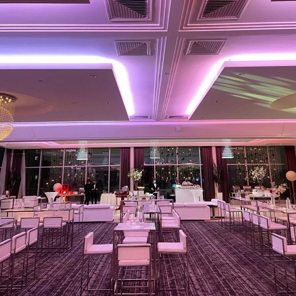 The image showcases an elegantly arranged interior of Le Crystal, a premier wedding venue listed on Mariage Elegant, Canada. The spacious room features a modern and sophisticated setting with soft purple lighting enhancing the ambiance. Sleek white chairs and tables are neatly organized, creating a welcoming and luxurious atmosphere for a wedding reception. The ceiling is adorned with recessed lighting, while large windows offer an open and airy feel. The decor includes floral arrangements and strategically placed lighting, highlighting the venue's versatility and elegance, making it an ideal choice for weddings.