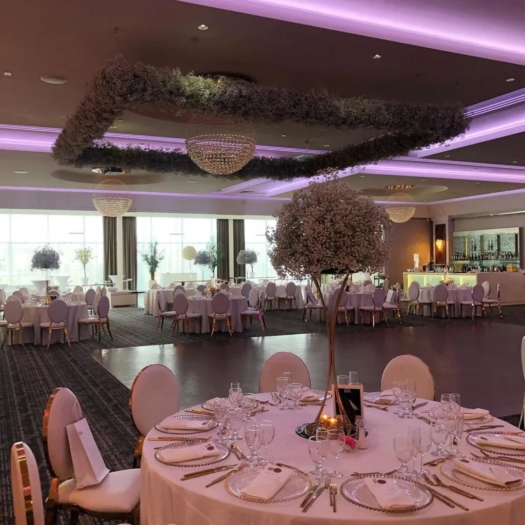 This image showcases the elegant interior of Le Crystal, a premier wedding venue in Canada, under the category of Wedding Venues on Mariage Elegant. The spacious hall features beautifully set round tables adorned with white tablecloths and neatly arranged glassware and cutlery. Each table is accented with tall centerpieces of white flowers. The ceiling is adorned with stunning chandeliers and suspended floral arrangements, creating a romantic ambiance. Soft purple lighting enhances the sophisticated atmosphere, making it an ideal setting for a memorable wedding celebration.