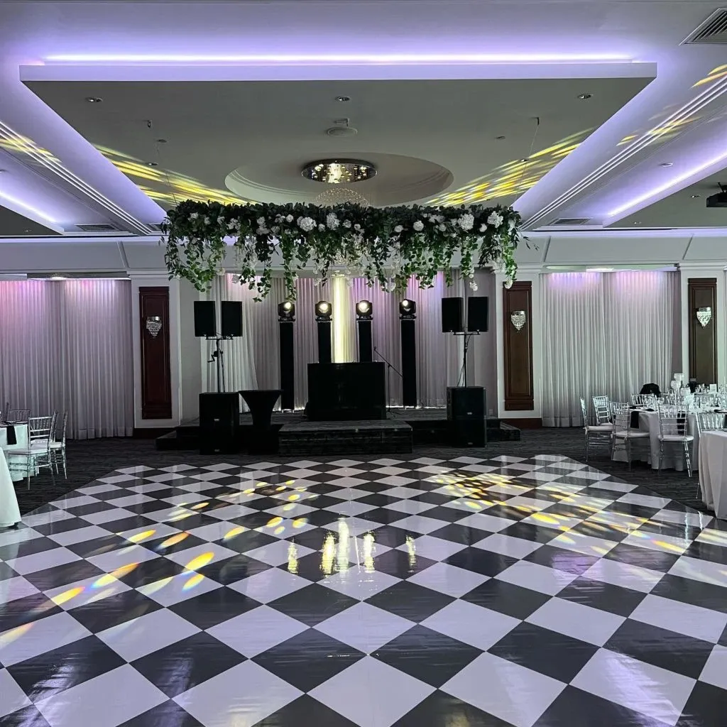 The image showcases a beautifully arranged wedding venue at Le Crystal, falling under the Wedding Venues category. The spacious hall features a striking black and white checkered dance floor illuminated by soft purple lighting. Elegant drapery adorns the walls, complemented by chandeliers providing a sophisticated atmosphere. A prominent floral arrangement with lush greenery hangs from the ceiling above the dance floor, adding a touch of natural elegance. In the background, a DJ booth is set up with speakers, ready for a wedding celebration. Tables and chairs are neatly arranged, indicating readiness for guests.