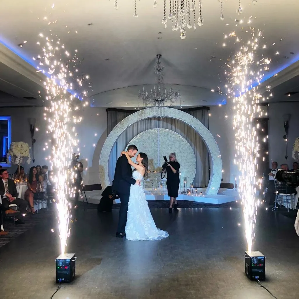 This image features a couple's first dance at a wedding hosted at Le Crystal, a premier wedding venue in Canada. The couple is elegantly dressed, with the bride in a beautiful white gown and the groom in a classic suit. They are surrounded by sparkling fireworks on either side, creating a magical ambiance. A stunning circular floral arch adorned with white flowers serves as a backdrop, enhancing the venue’s sophisticated decor. Guests are seated around the dance floor, witnessing the special moment. The elegant chandeliers and ambient blue lighting add to the venue's luxurious atmosphere, highlighting its suitability for an unforgettable wedding celebration.