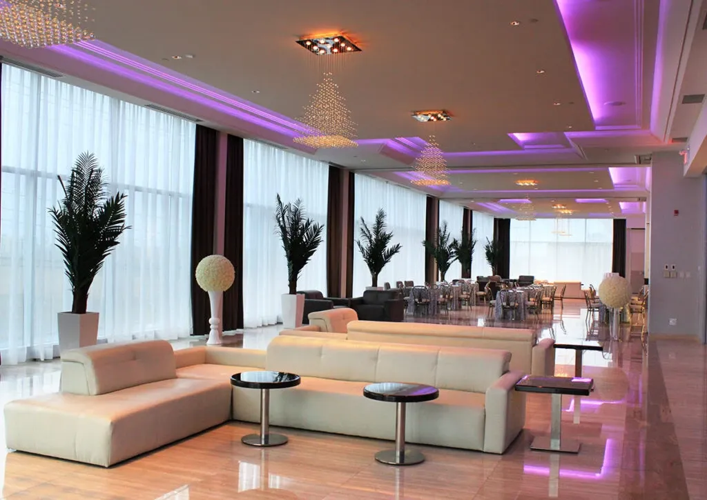 The image showcases the elegant interior of Le Crystal, a venue in the Wedding Venues category, featured on Mariage Elegant, Canada. The spacious room is designed with modern luxury in mind, featuring a series of long, white couches paired with sleek, round tables. The ceiling is adorned with sparkling chandeliers and enhanced by soft purple ambient lighting, creating a romantic atmosphere. Large windows with sheer curtains allow natural light to fill the space, while tall green plants add a touch of nature to the decor. The setting is ideal for hosting sophisticated wedding receptions or events.