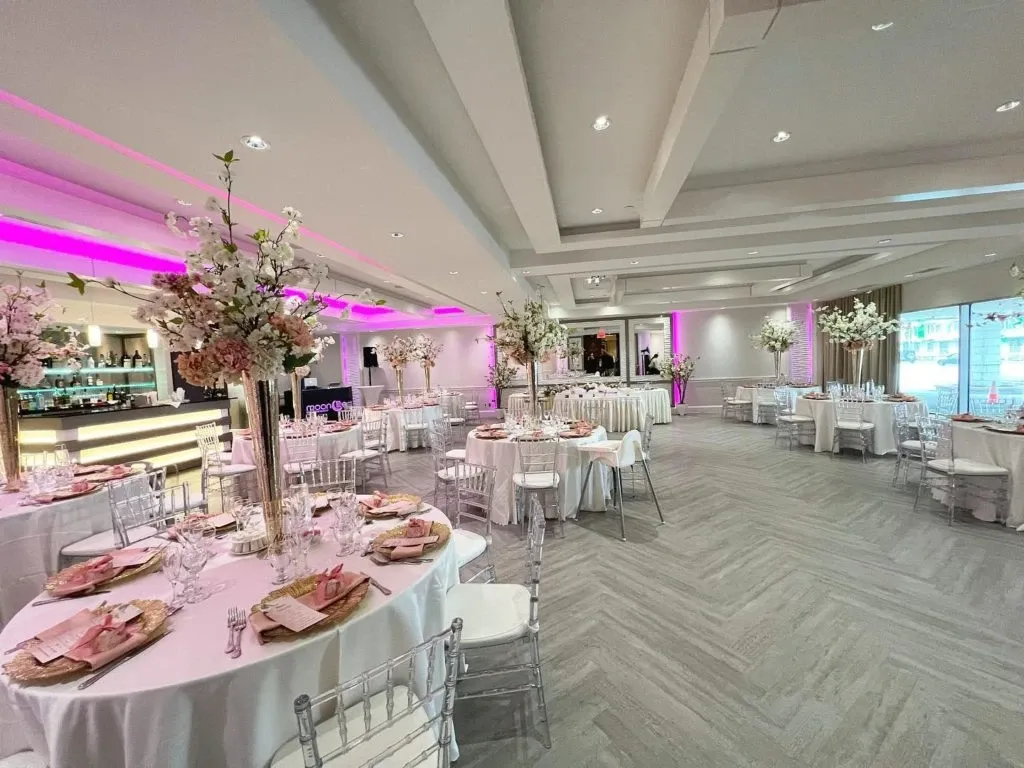 The image showcases a beautifully decorated wedding venue at Le Crystal, categorized under Wedding Venues on Mariage Elegant, Canada. The spacious room features elegantly set tables adorned with tall floral centerpieces, enhancing the romantic ambiance. The soft, pink uplighting and modern ceiling design create a luxurious atmosphere, suitable for a memorable wedding reception. The venue also includes a well-stocked bar in the background, adding to the sophisticated setting designed for celebrating special occasions.